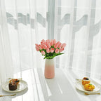 Pink tulips in a vase on a table with pastries, against a white curtain background