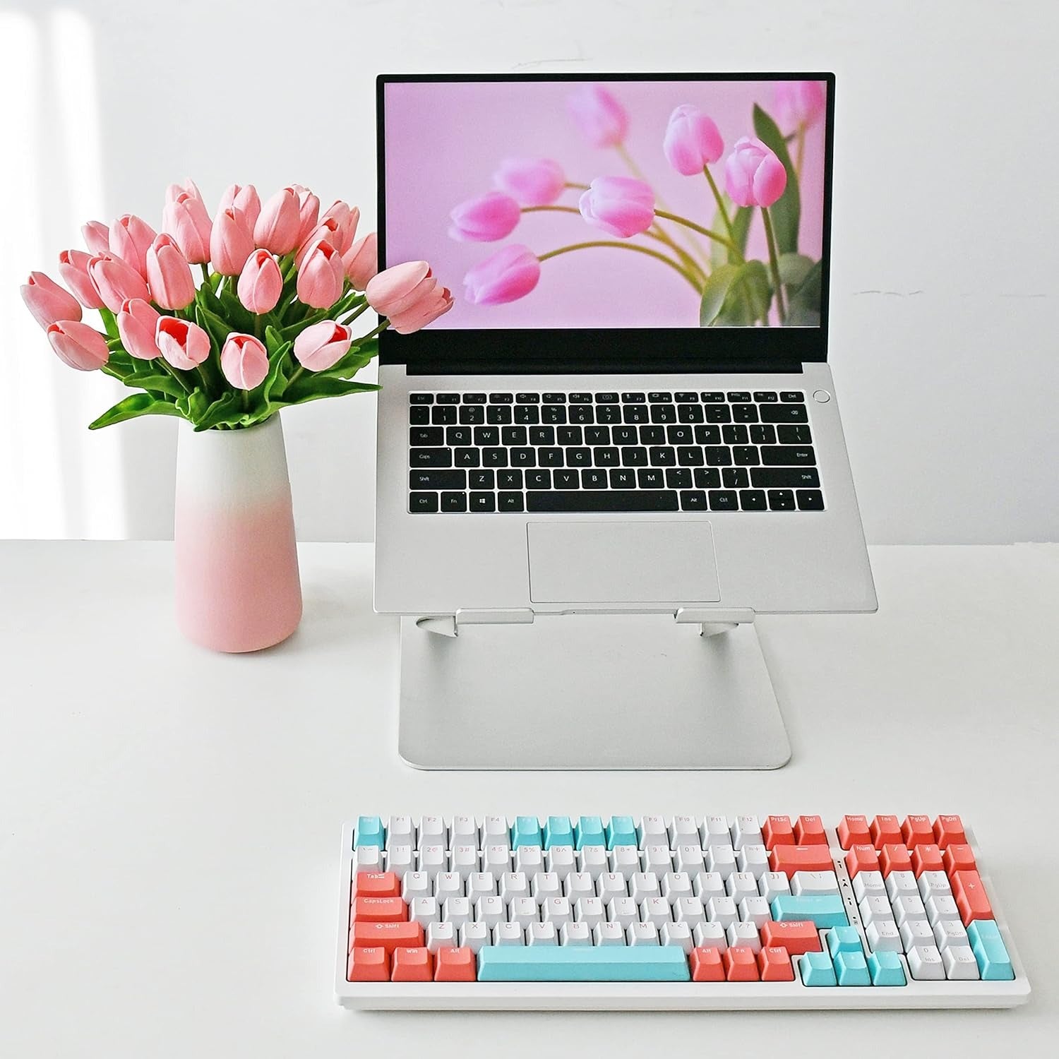Laptop with a floral display on screen, next to a vase of pink tulips and a colorful keyboard on a white surface.