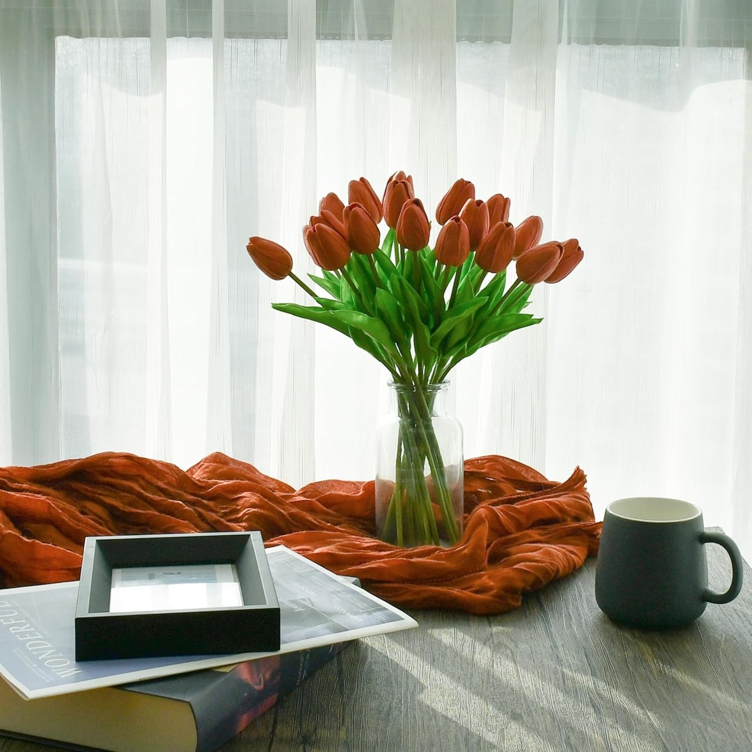 Bouquet of red tulips in a vase on a table with a mug and books, against a white curtain background.