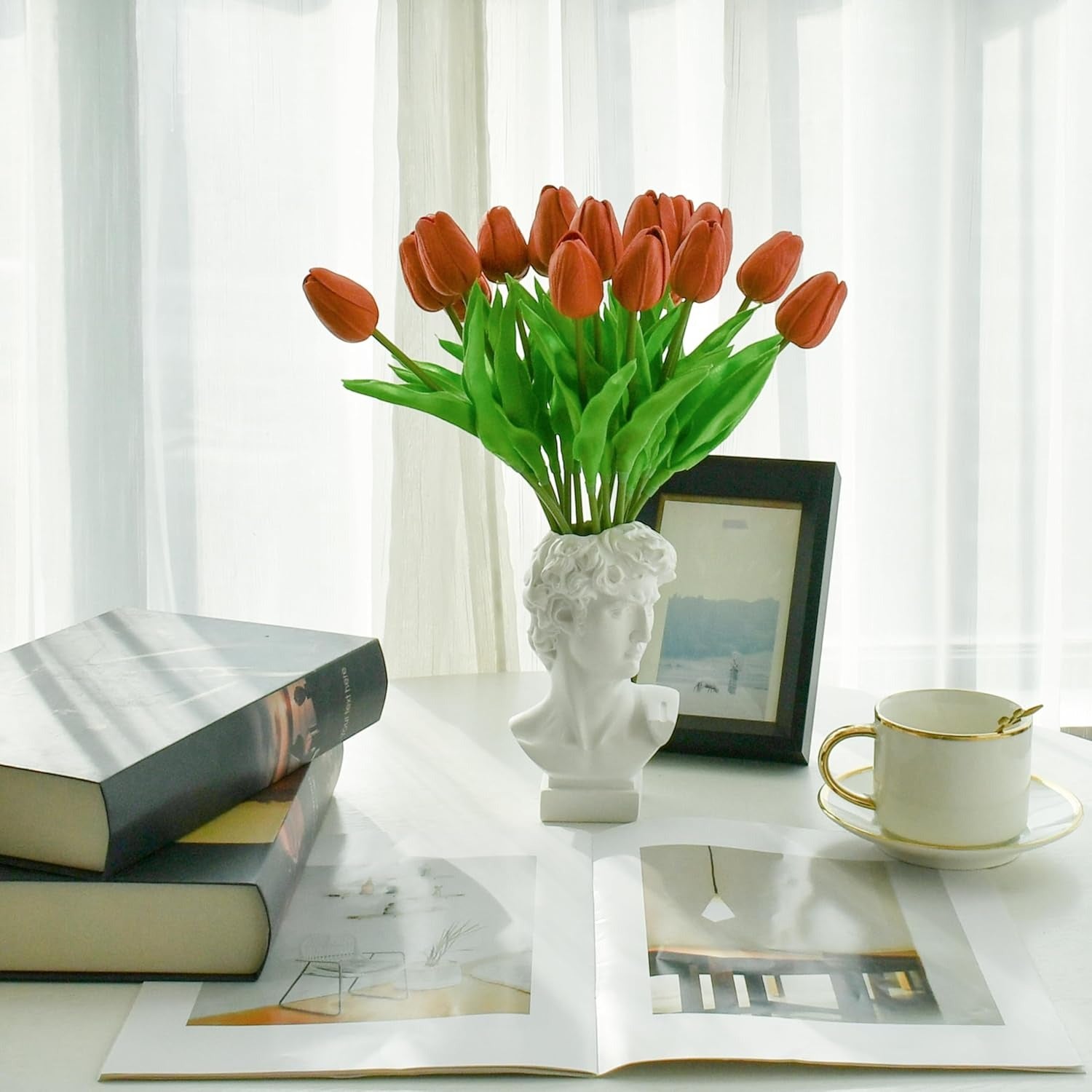 White vase with red tulips on a table with books and a cup.