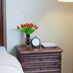 Wooden nightstand with a vase of orange tulips, clock, and square plate on a blurred bedroom background.