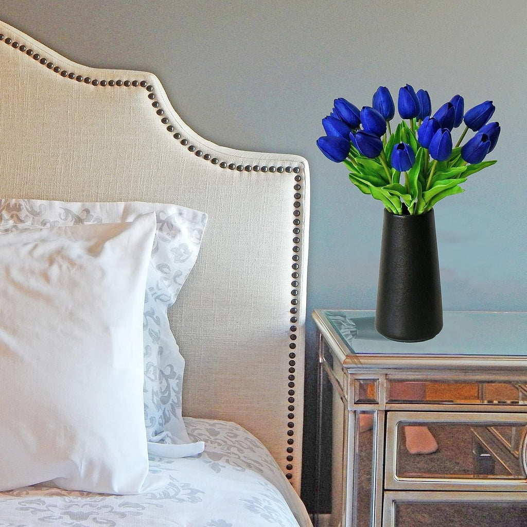 Vase with blue tulips on a nightstand next to a bed with white pillows.