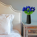 Vase with blue tulips on a nightstand next to a bed with white pillows.