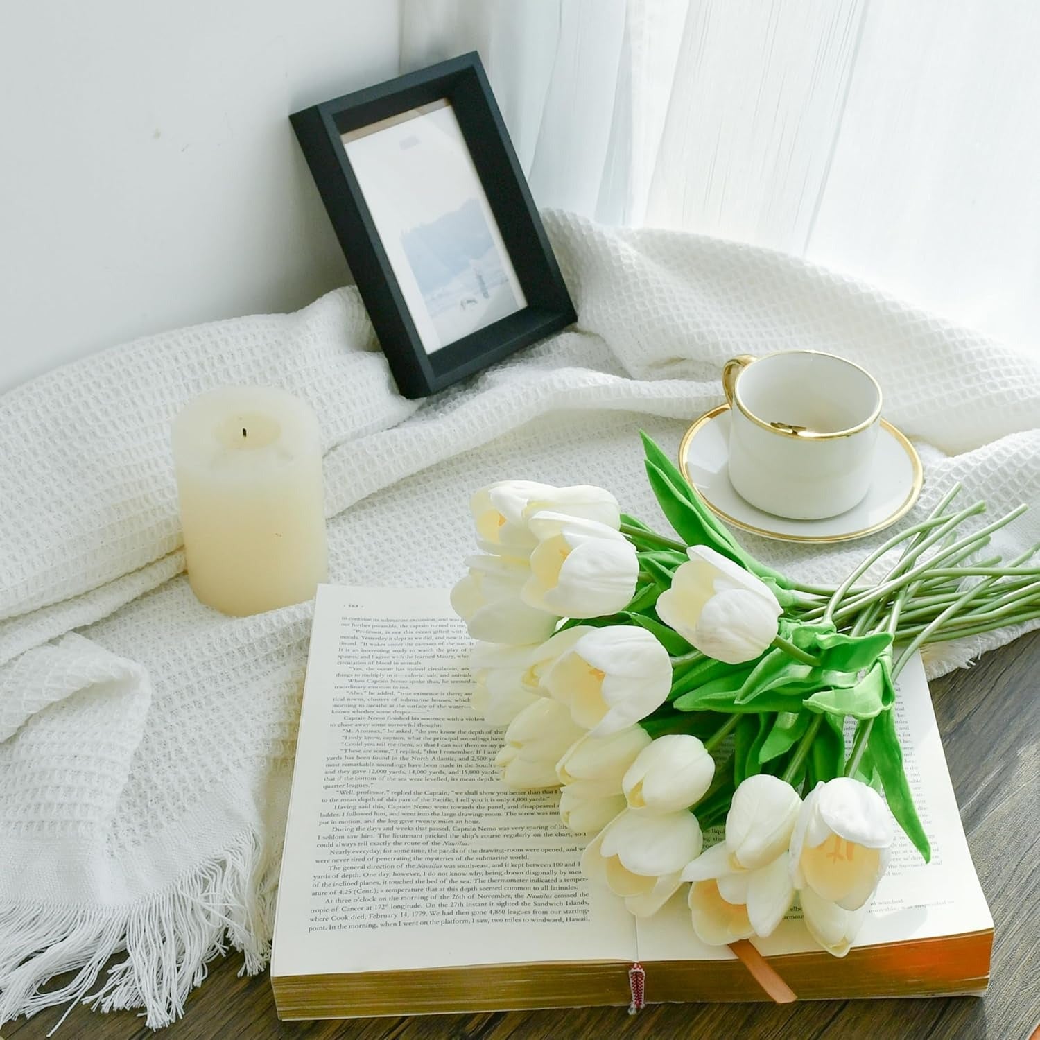 White tulips on an open book with a cup, saucer, and candle on a white blanket.