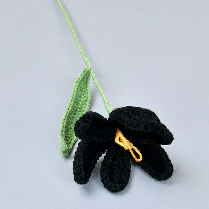 Crocheted black flower with a yellow center and green leaf on a light gray background