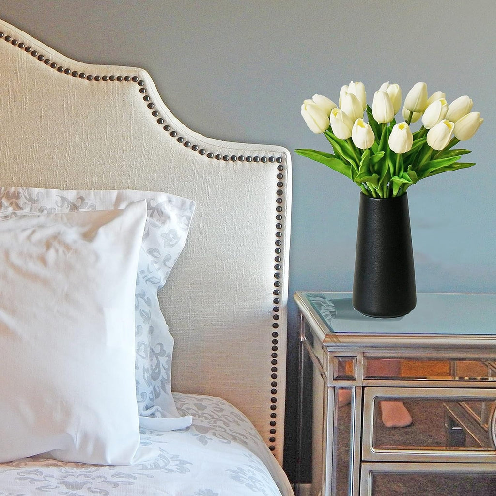 White tulips in a black vase on a nightstand next to a bed with white pillows.