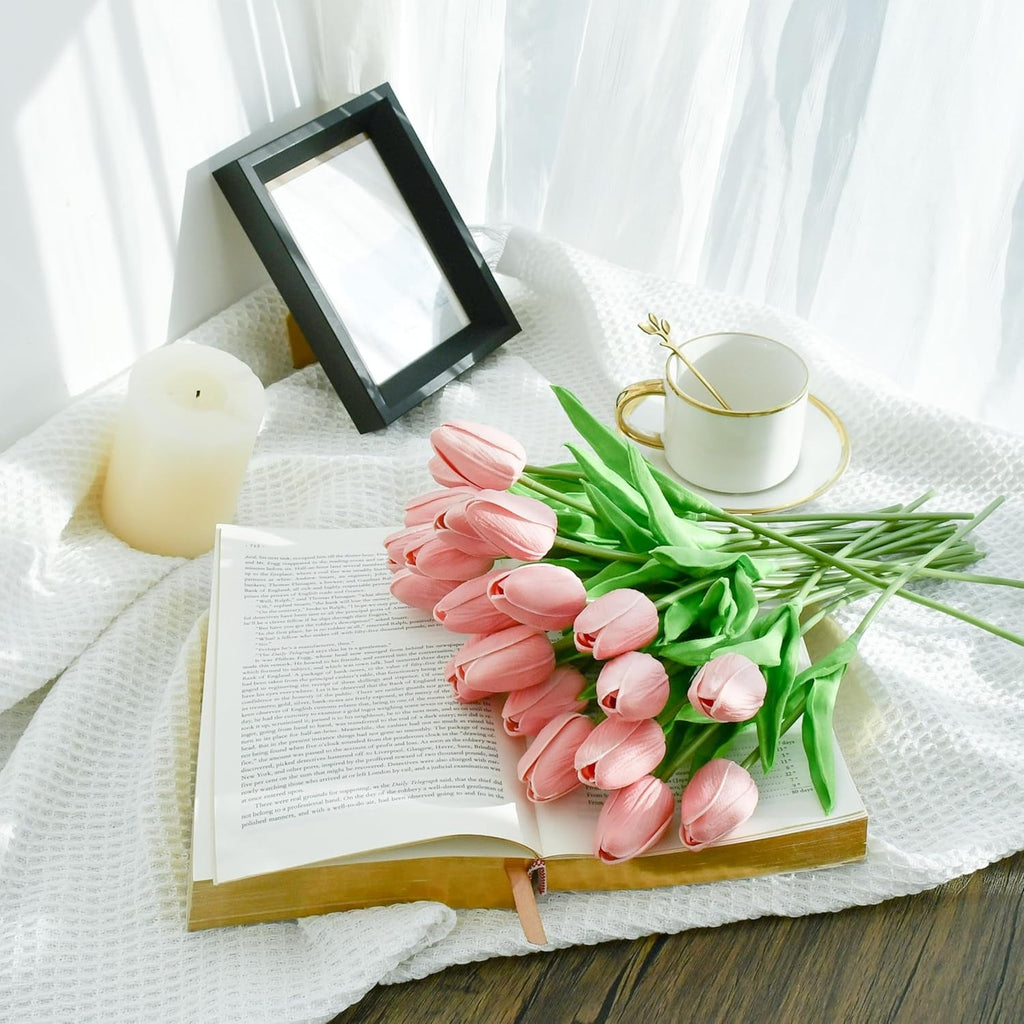Pink tulips on an open book with a cup, saucer, and candle on a white cloth.