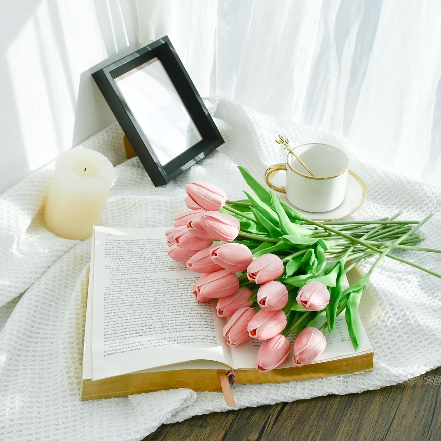 Pink tulips on an open book with a cup, saucer, and candle on a white cloth.