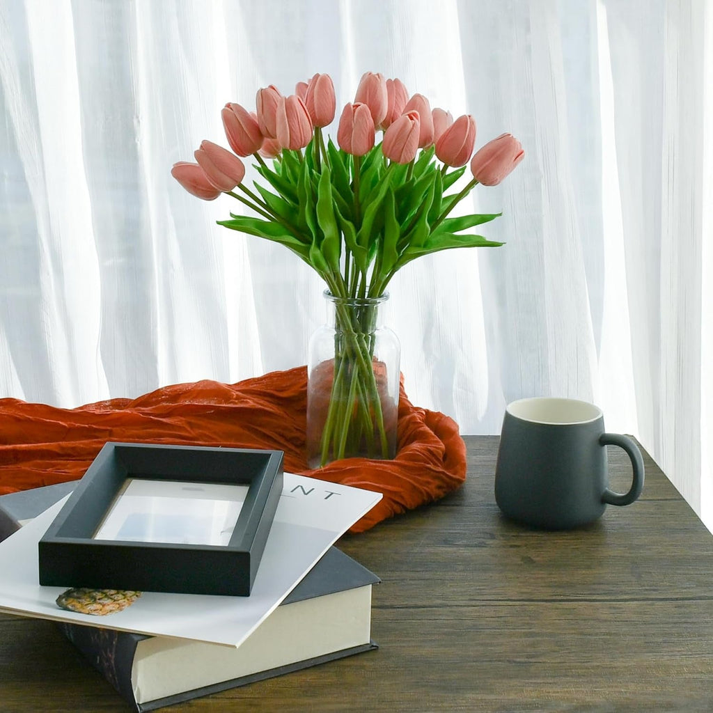 Vase of pink tulips on a wooden table with a mug and books underneath, against a white curtain background.