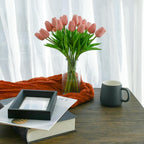 Vase of pink tulips on a wooden table with a mug and books underneath, against a white curtain background.