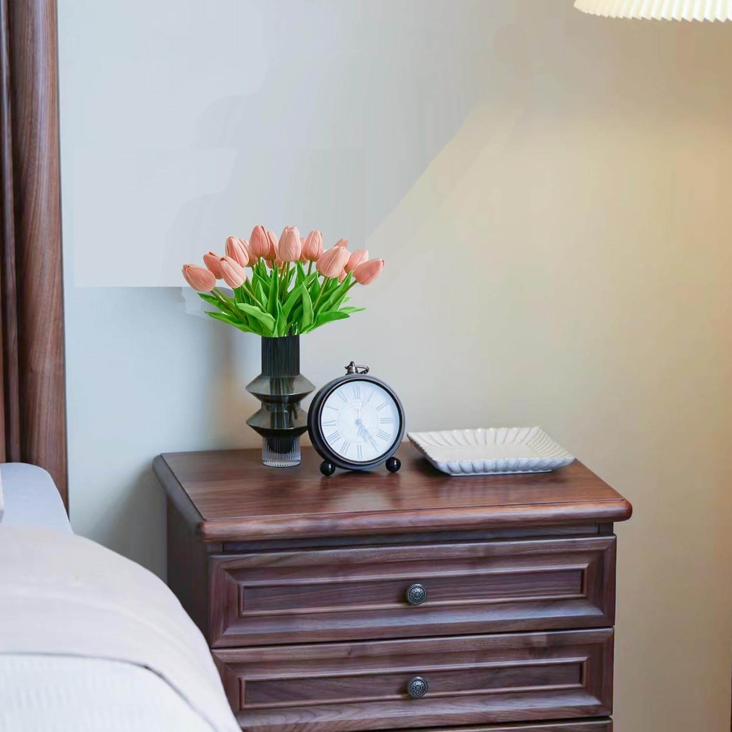 Wooden nightstand with a vase of pink tulips, clock, and square plate on a blurred bedroom background.