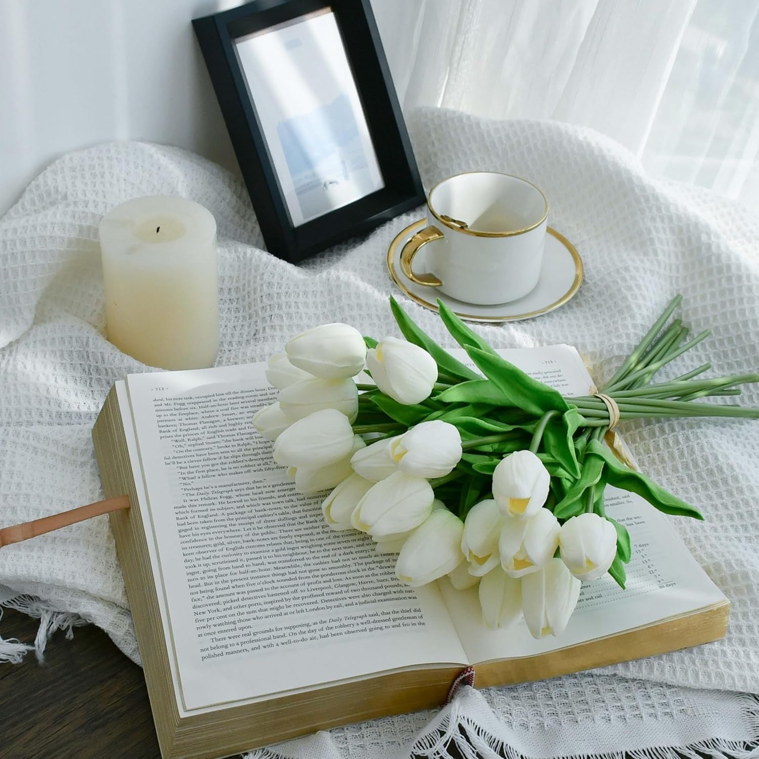 Open book with white tulips, a candle, and a cup on a white fabric background