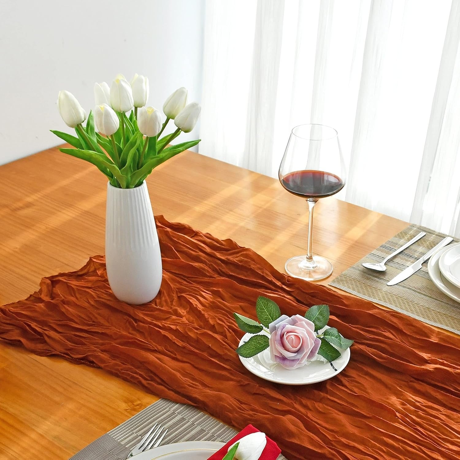 Table setting with a vase of white tulips, a glass of red wine, and a small plate with a pink flower on a wooden table.