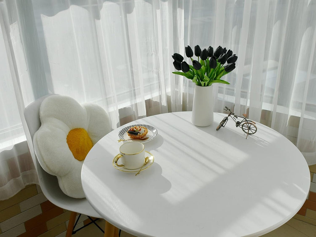 Round white table with a vase of black tulips, a cup, and a decorative item in a room with white curtains.