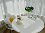 Round white table with a vase of black tulips, a cup, and a decorative item in a room with white curtains.