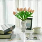 Bouquet of pink tulips in a white vase on a table with books and a cup.