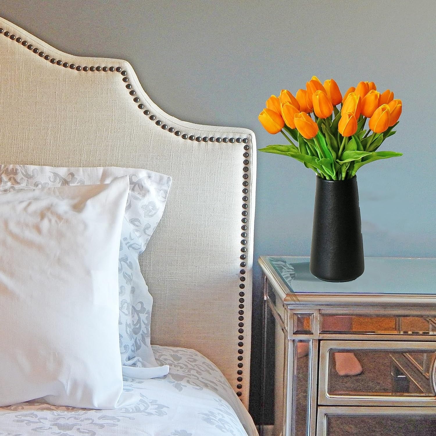 Orange tulips in a black vase on a nightstand next to a bed with white pillows.