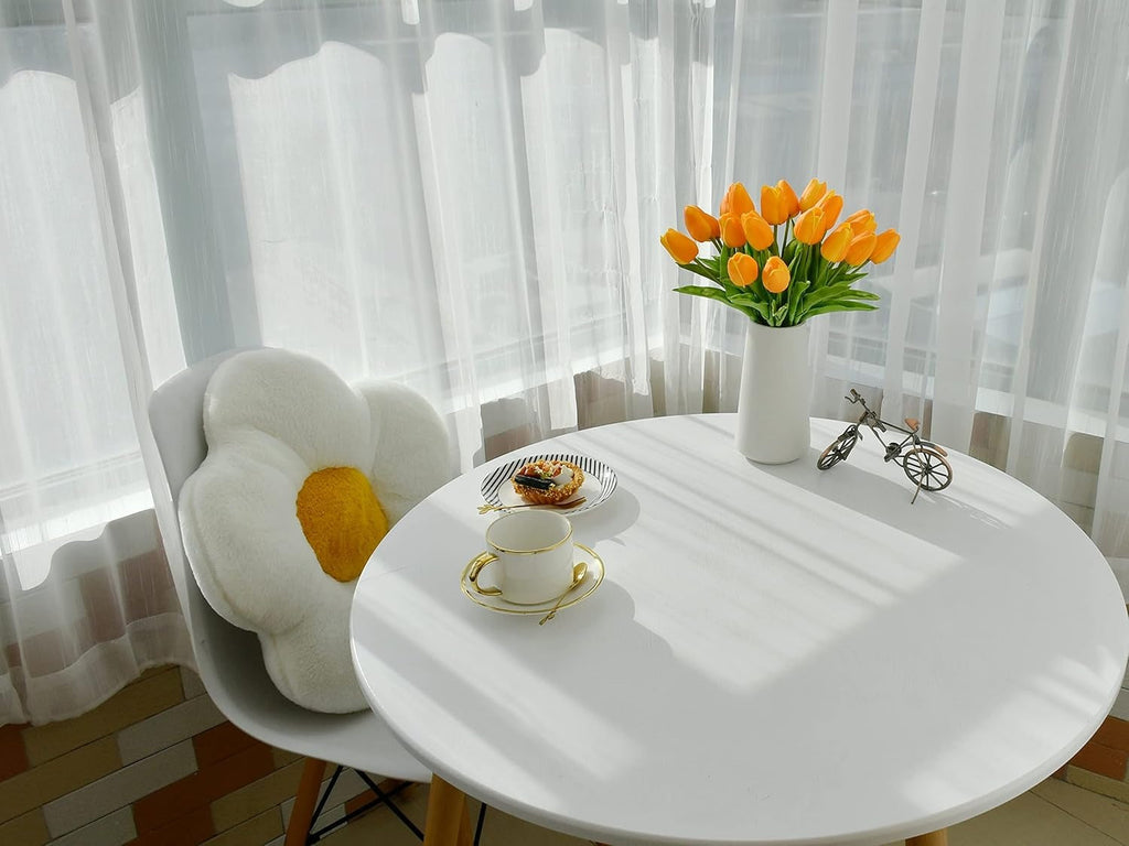 Round white table with a vase of yellow tulips and a teacup, set against a bright window with white curtains.