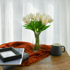 Vase of white tulips on a wooden table with a mug and books in front of sheer curtains.