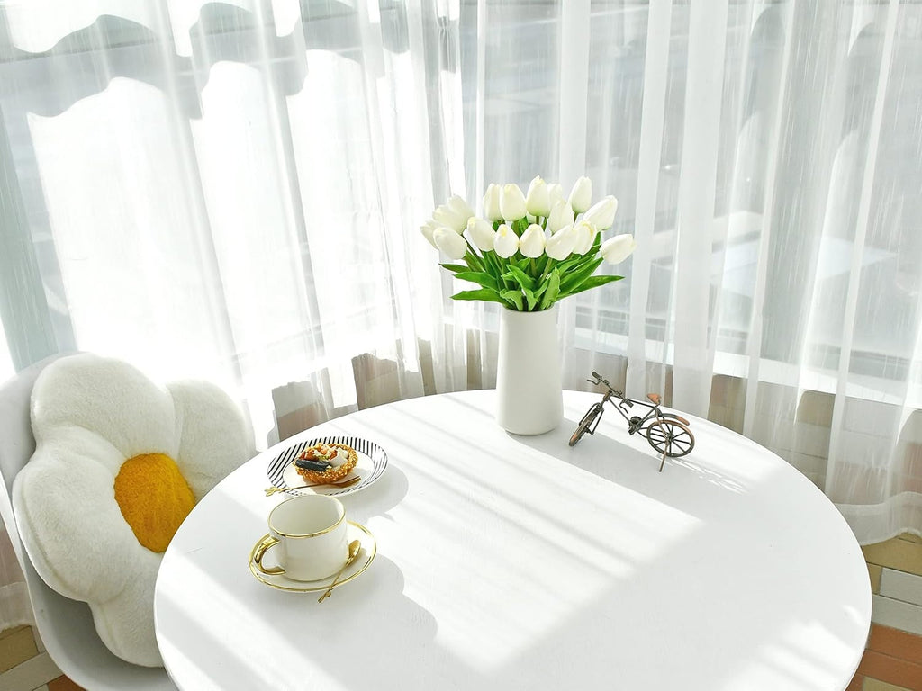 White table with a vase of white tulips, a cup, and a small bicycle decoration in front of sheer curtains.