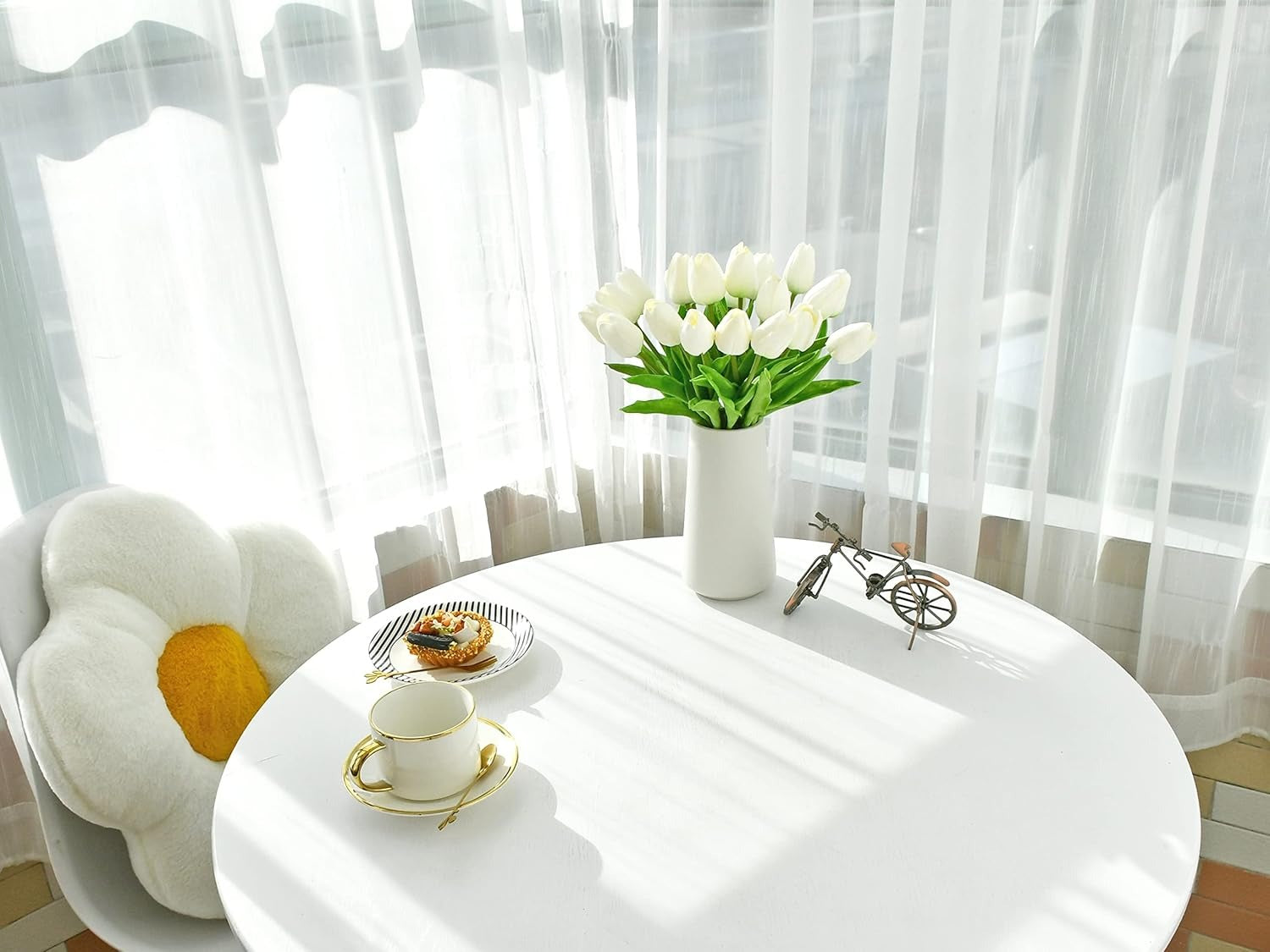 White table with a vase of white tulips, a cup, and a small bicycle decoration in front of sheer curtains.
