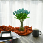 Vase with blue tulips on a table with an orange scarf, black mug, and photo frame.