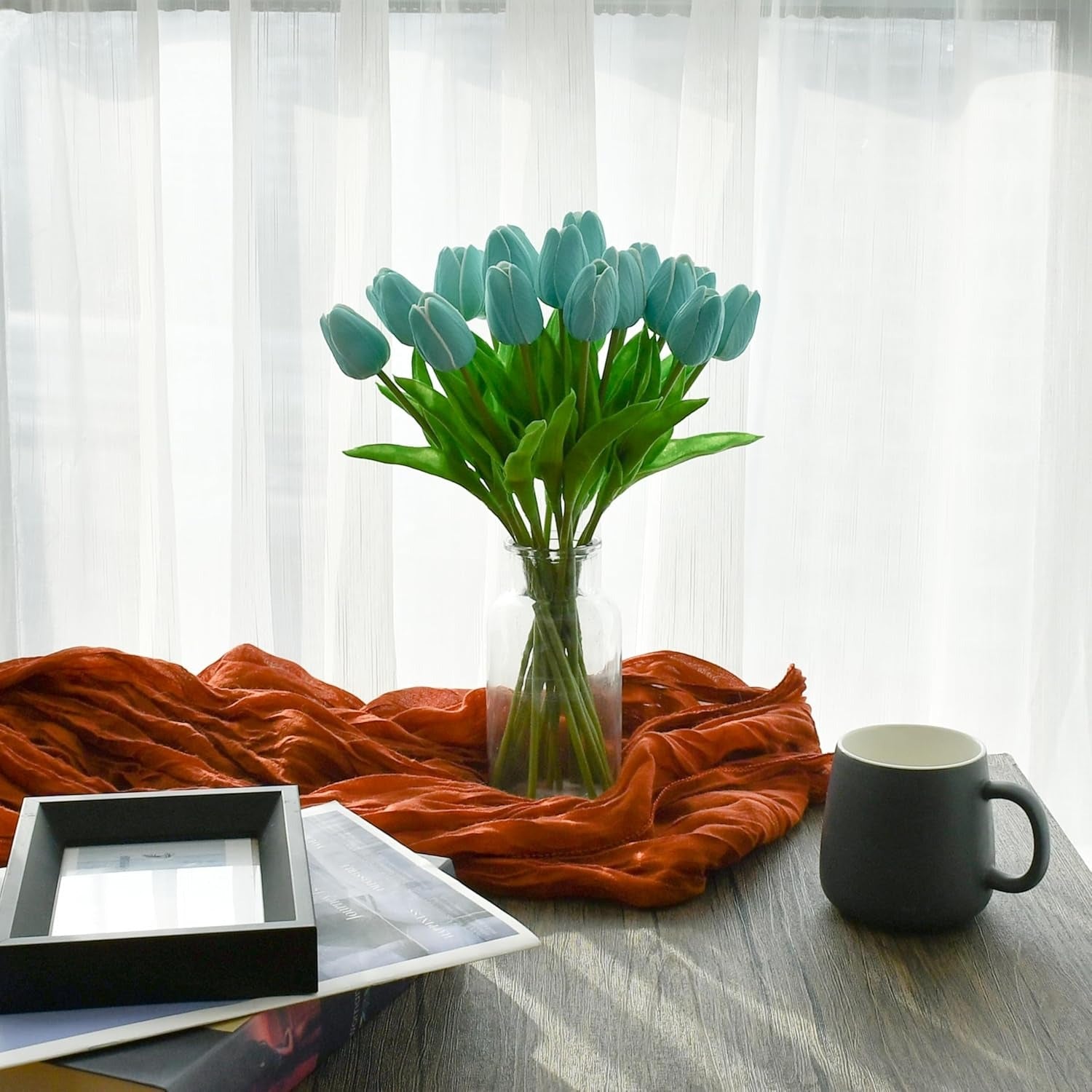 Vase with blue tulips on a table with an orange scarf, black mug, and photo frame.