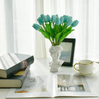 Teal tulips in a white vase on a table with books and a cup.